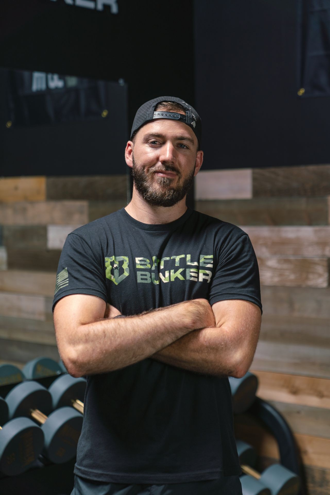 Man in a black t-shirt and cap standing with arms crossed in front of gym equipment.